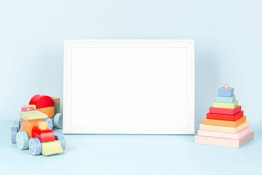 Toys background. Empty white wooden picture frame with blank mock up copy space standing next to wooden train and colorful toy pyramid on pastel blue background. Front view