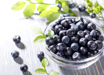 Sun backlit glass bowl with ripe fresh blueberries stands on grey wooden table close-up. Natural superfood.