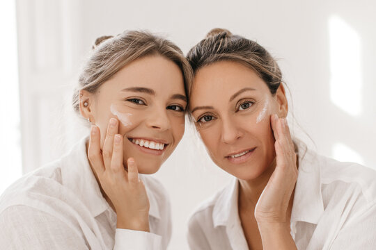Close-up of two attractive young women looking at camera applying cream to their face on white background. Blonde ladies in casual wear. Beauty needs care concept