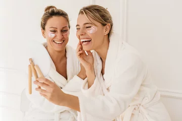Abwaschbare Fototapete Spa Cheerful fair-skinned young women wear terry dressing gowns laugh looking in mirror on white background. Blondes use hydrogel patches under their eyes. Home spa facial concept