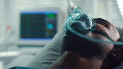 Portrait patient breathing oxygen mask in ward. Heartbeat monitor at window.
