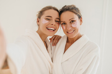 Happy caucasian mother with daughter wearing hydrogel patches smiling looking at camera on white background. Blondes wear robes to take care of their skin. Cosmetic natural trend. - obrazy, fototapety, plakaty