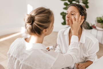 Obraz na płótnie Canvas Caucasian girl make-up artist applies cream on face of beautiful adult woman sitting in white room. Blonde ladies wear casual shirts. Skin care and hydration concept