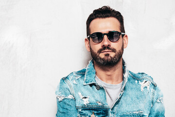 Fototapeta premium Portrait of handsome confident stylish hipster lambersexual model. Sexy man dressed in jeans jacket. Fashion male isolated on grey wall in studio. In sunglasses