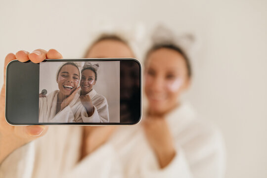 Attractive young smiling caucasian women make photo on smartphone on white background. Girls wear robes, headbands and use patches. Female beauty rituals, skin care cosmetic products concept