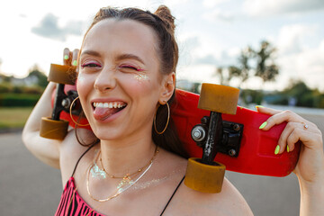 Positive young teenage female skater with skateboard showing tongue and making funny face while looking at camera in city street against sunset sky in summer  - obrazy, fototapety, plakaty