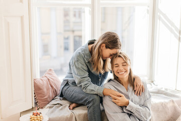 Obraz premium Young caucasian girl with closed eyes and her mother are resting together at window on sunny warm day. Blondes wear shirts and jeans. Concept of relationships and sharing time.