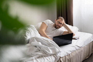 smiling man using Tablet Computer is lying on a bed in the morning. work from home. freelancer.