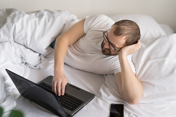 Naklejka premium smiling man using Tablet Computer is lying on a bed in the morning. work from home. freelancer. social networks concept. man chatting with friends.