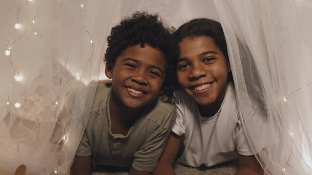 Chest-up portrait of cute African American brother and sister lying in illuminated with garland blanket fort, looking and smiling on camera