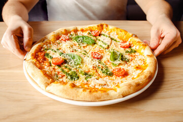 Italian pizza with chicken, cherry tomatoes, mozzarella and parmesan cheese, basil on plate on wooden table and male caucasian hands. Top front view, lifestyle photo, closeup.