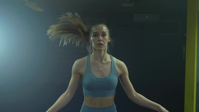 Zoom-in view of a young fit Caucasian woman in sportswear, skipping a rope, during fitness training in an empty modern health club, handheld shot.