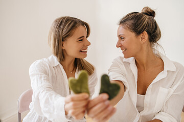Positive young caucasian blonde pointing at camera with face scraper on white background. Women wear shirts and look at each other. Concept of natural cosmetics, wrinkle smoothing