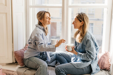 Two young caucasian female friends on coffee break spend time together sitting on windowsill day. Blondes wear jeans and shirts. People, communication and friendship concept