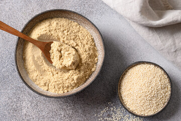 Sesame flour in bowl and white sesame seeds on gray background for cooking gluten-free and low carbohydrate dessert. Sesame flour includes ample minerals, natural antioxidants and vitamins.