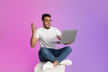 Excited young Arab man sitting cross legged with laptop, feeling overjoyed over huge online sale in neon light