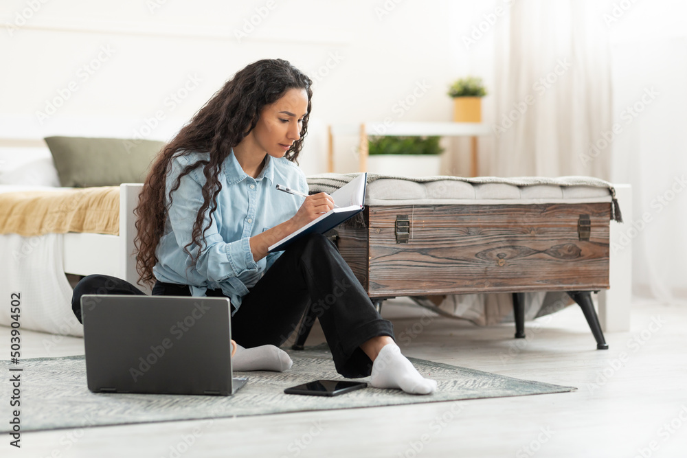 Poster woman sitting on floor, using computer and writing in notebook