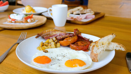 American breakfast with sunny side fried eggs, bacon, tomatoes, bread toasts, baked potatoes and coffee cup on wooden table in restaurant. Delicious served lunch or brunch food set meals in cafe.