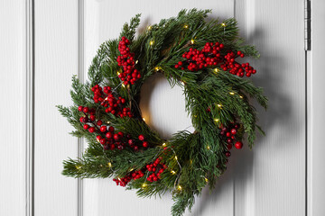 Beautiful Christmas wreath with red berries and fairy lights hanging on white door