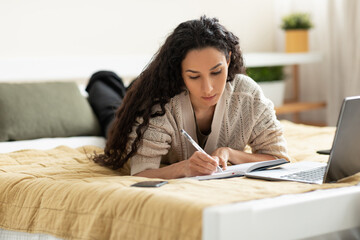 Woman lying on bed, using computer and writing in notebook - obrazy, fototapety, plakaty