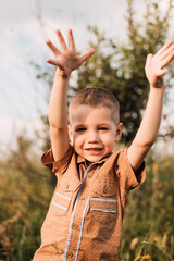 A little boy stands in a village in a field, smiling and raising his hands up.