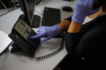 An employee in the office makes a call on a landline phone wearing gloves. Phone close-up. In gray tones. Protection, virus.Stationary phone and headset on desk indoors, closeup. 