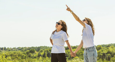 Two young woman wearing sunglasses holds hands in nature, pointing finger at the sky.