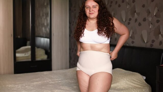Curly overweight pleased woman in white underwear stand near bed in bedroom and pull slimming panties over sagging stomach. Fat burning treatment of thick cellulite body. Weight loss control.