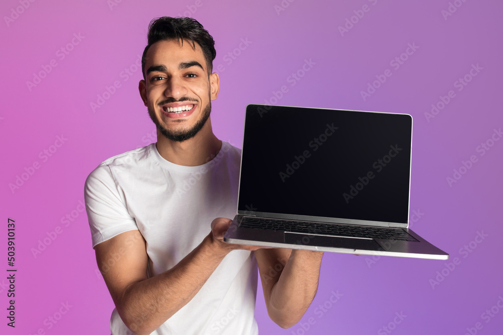 Poster Smiling millennial Arab man showing laptop computer with empty black screen in neon light, mockup for website or ad