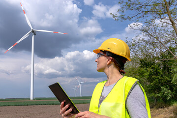 A female engineer standing on a field, looking at windmills on a wind farm, and making notes on a digital tablet. Green renewable energy concept.