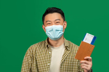 Happy mature asian man in medical mask holding plane tickets and international passport, posing on green background