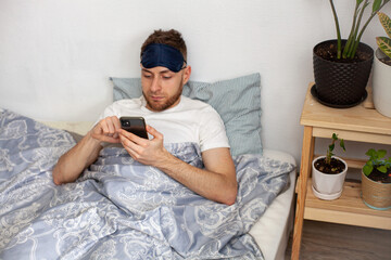 young man lying in bed in the morning wearing a sleep mask and checking social media