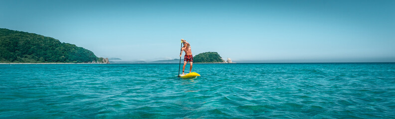 Tanned man stands on a yellow paddle board and paddles. Active sport on the beach in the turquoise...