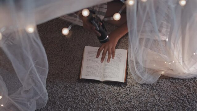 Top view of unrecognizable child sitting on floor in handmade tent at home, reading book using flashlight at night