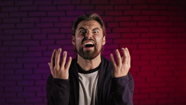 Portrait of aggressive bearded man shouting angry and gesturing emotionally in studio with neon light. Caucasian male person feeling furious indoors.