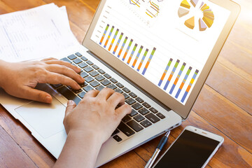 Close-up business woman working in an office interior on a computer holding a smartphone and looking at a diagrammed screen. Office worker using mobile phone and laptop.