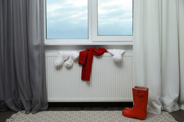 Obraz premium Heating radiator with hat, socks, scarf and rubber boots near window indoors