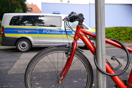 Ein rotes Fahrrad steht angeschlossen an einem Stra&szlig;enschild, w&auml;hrend im Hintergrund ein Polizeiauto parkt.