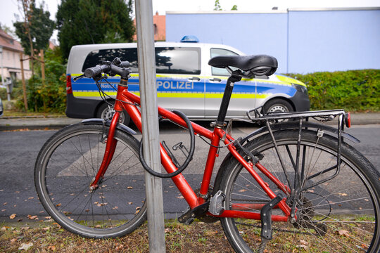 Ein rotes Fahrrad steht angeschlossen an einem Stra&szlig;enschild, w&auml;hrend im Hintergrund ein Polizeiauto parkt.