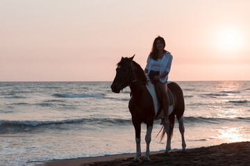 Fototapeta na wymiar Woman in summer clothes enjoys riding a horse on a beautiful sandy beach at sunset. Selective focus 
