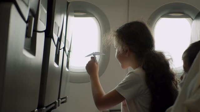Child sitting by aircraft window and playing with little paper plane, during flight on airplane. Child's hand with small paper plane against the background of airplane window.