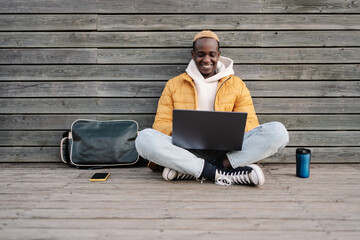 African American male university student sitting outdoors working on laptop smiling wearing yellow...