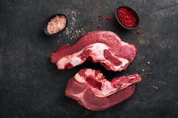 Raw beef meat. Rough piece of meat on bone for roast or soup with salt, pepper, thyme and rosemary on a black concrete background. Entrecote. Raw cowboy steak. Top view.