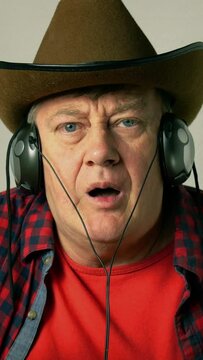 Expressive adult senior man in a cowboy hat, listens to music on his headphones.