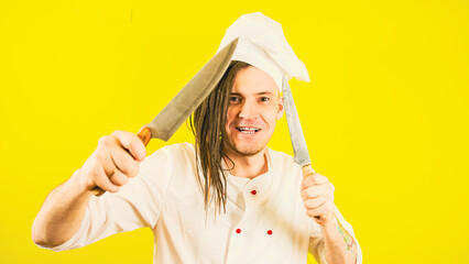 Young insane man dressed as chef with knives on yellow background. Male cook in white hat and shirt posing with kitchen knives.