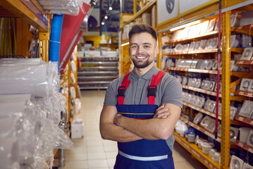 Confident friendly man working in warehouse in hypermarket of building materials. Smiling young male worker in coveralls standing with folded arms in one of aisles between shelves with goods. - obrazy, fototapety, plakaty