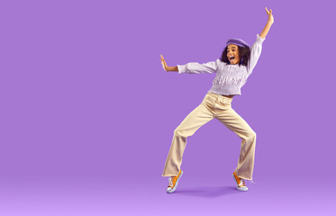 Overjoyed African American teen girl in fashionable clothes dance on violet studio background. Funny black teenage young woman have fun make dancer moves. Hobby and entertainment.