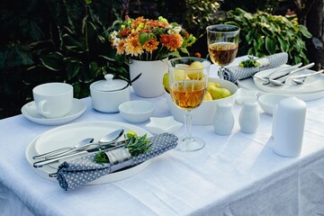 Table set for outdoor eating on a table in the garden. Elegant table setting with white dishes on a white linen tablecloth. Sunny day light. Copy space