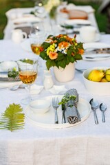 Elegant table setting with white dishes on a white linen tablecloth. Fruits, drinks in crystal glasses. Summer holiday table set up. Outdoor eating on a table in the garden. Vertical shot