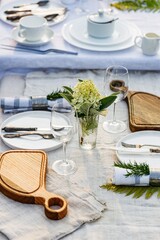 Summer holiday table set up. Elegant table setting with white dishes on a white linen tablecloth. Table set for outdoor eating in the garden. Vertical shot